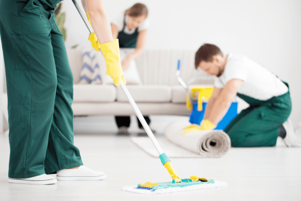 three professional rug cleaners in picture, a girl behind cleaning sofa, 2nd person is unrolling an area rug and third person is mopping the floor, seems like professional cleaning is in progress.