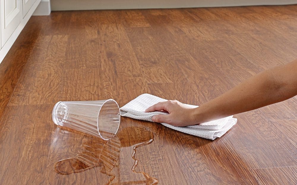 a glass of water spilled on vinyl floor and a person is cleaning water with a white cloth.